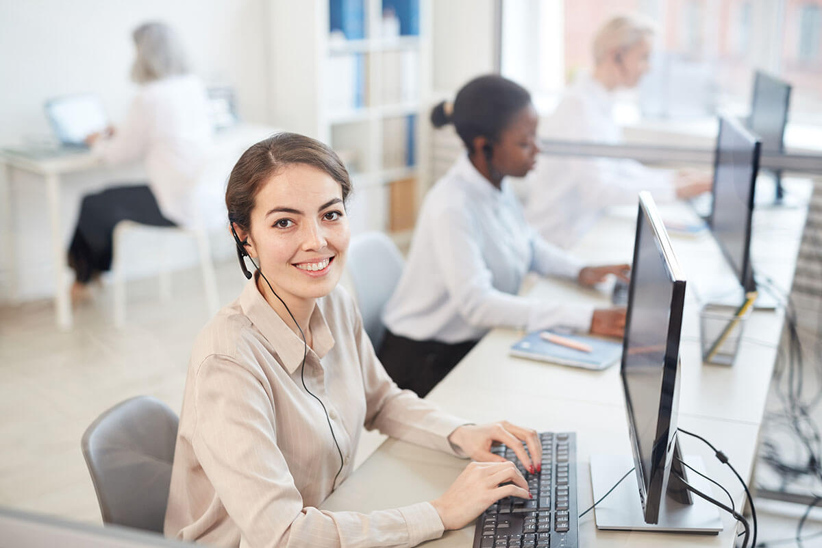 High angle portrait of female operator wearing headset and smiling at camera while sitting in row at call center.