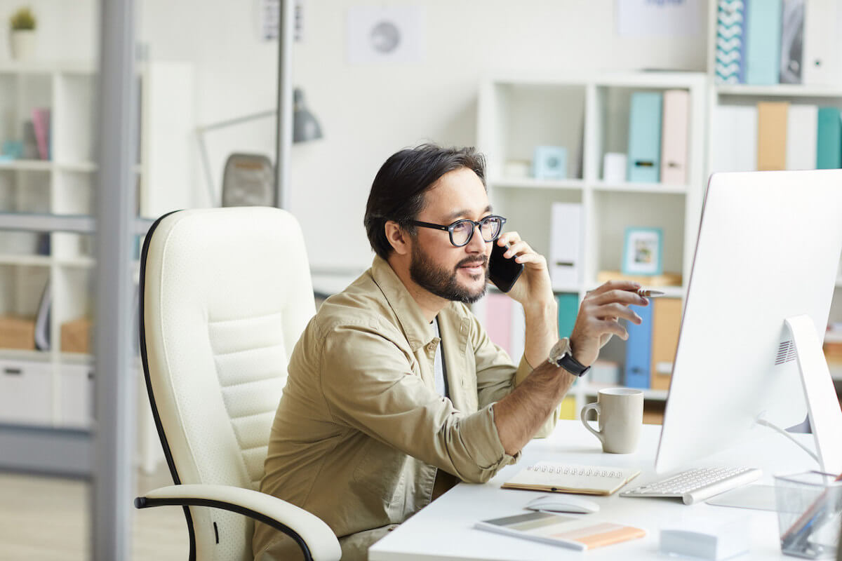 Smart young asian programmer in casual shirt sitting at table with notepads and pointing at computer